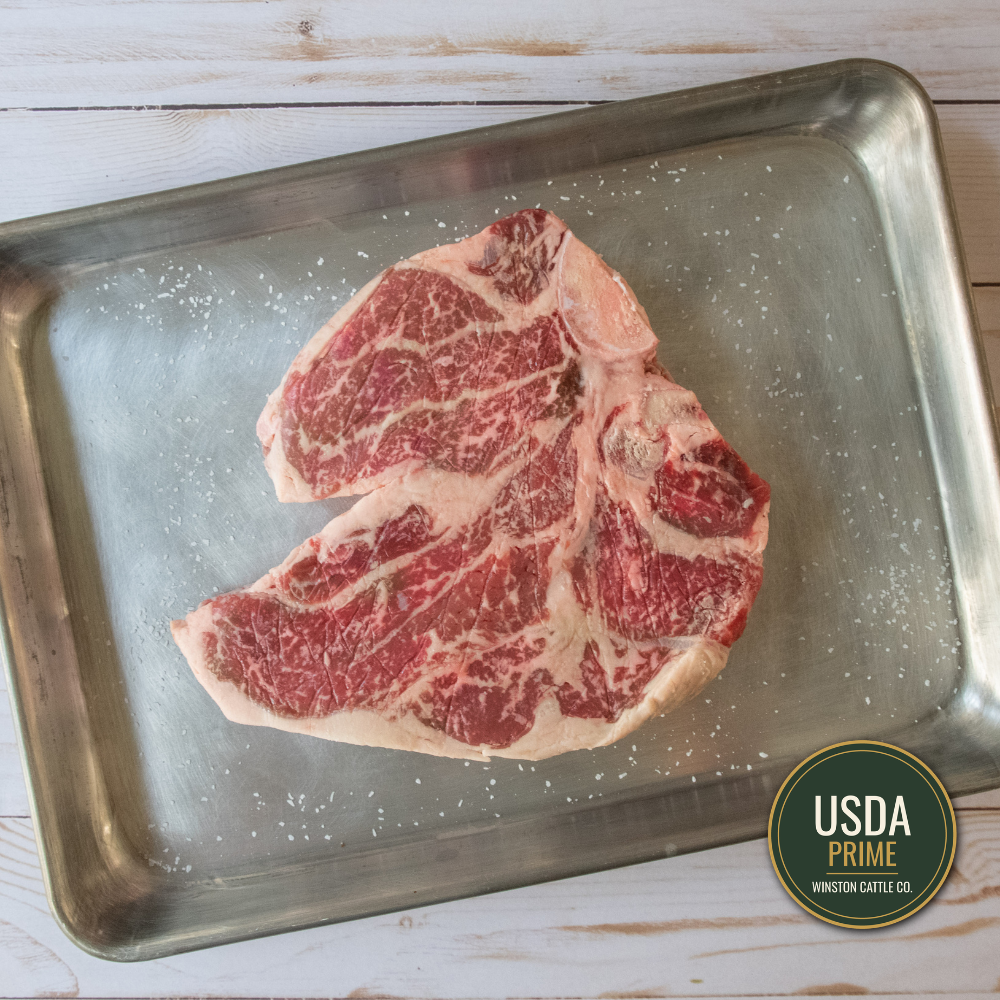 A USDA Prime T-bone steak with marbled fat, resting on a stainless steel baking sheet sprinkled with coarse sea salt, set on a white wooden surface. A USDA Prime Winston-Slate Co.