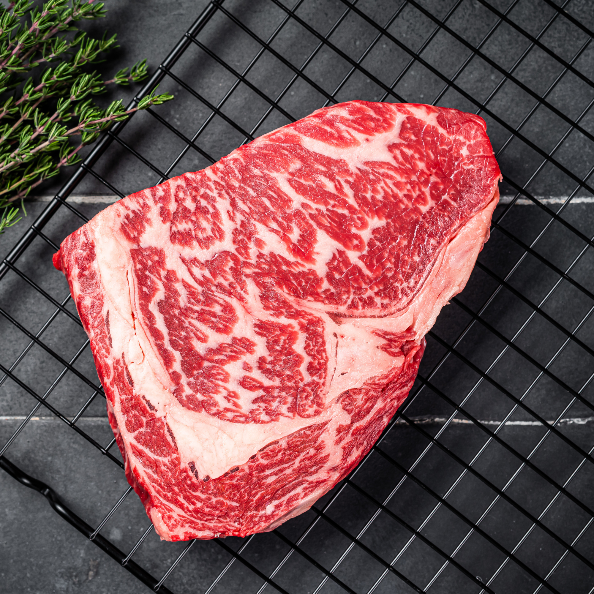 A raw, thickly cut 100% Fullblood American Wagyu ribeye steak resting on a black wire cooling tray, showcasing elite Japanese bloodline intramuscular snowflake marbling at Winston Cattle Co.
