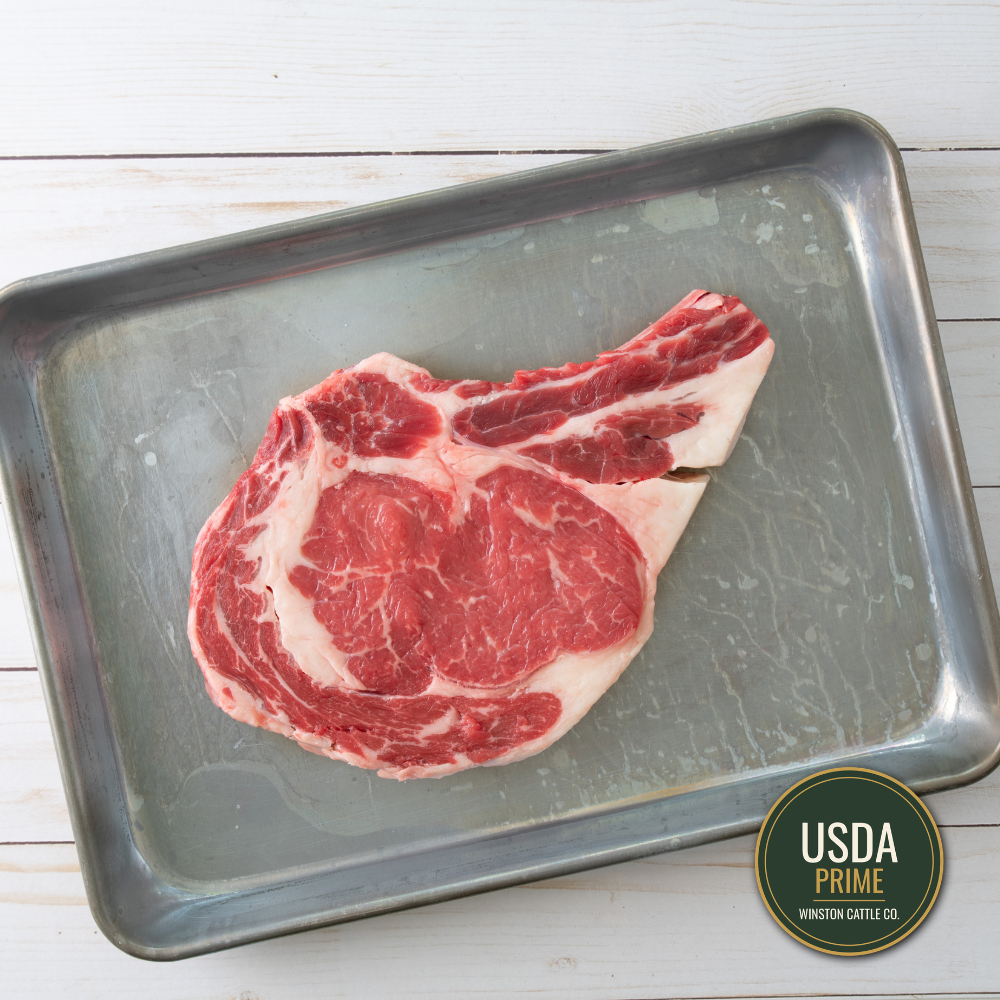 Prime USDA-graded Bone-in Ribeye Cowboy steak with marbling, presented on a metal baking tray over a white wood background.