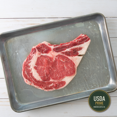 Prime USDA-graded Bone-in Ribeye Cowboy steak with marbling, presented on a metal baking tray over a white wood background.