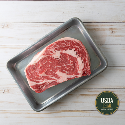 A USDA Prime boneless Ribeye steak with marbling, presented on a rectangular metal tray over a white wood plank background, with a USDA Prime logo badge in the bottom right corner. Winston Cattle Co.