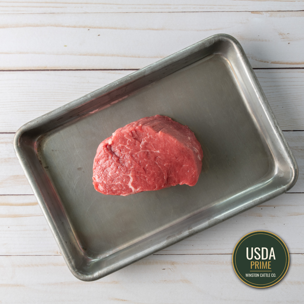 A usda prime filet mignon beef cut, likely a tenderloin or filet mignon, sits on a rectangular metal baking tray over a white wood plank background. A USDA Prime Winston Cattle Co.