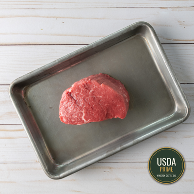 A usda prime filet mignon beef cut, likely a tenderloin or filet mignon, sits on a rectangular metal baking tray over a white wood plank background. A USDA Prime Winston Cattle Co.