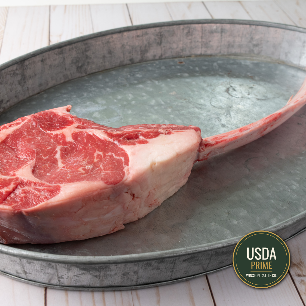 A large USDA Tomahawk ribeye steak with bone lies in a galvanized metal tray on a white wood surface. A USDA Prime Winston Cattle Co.