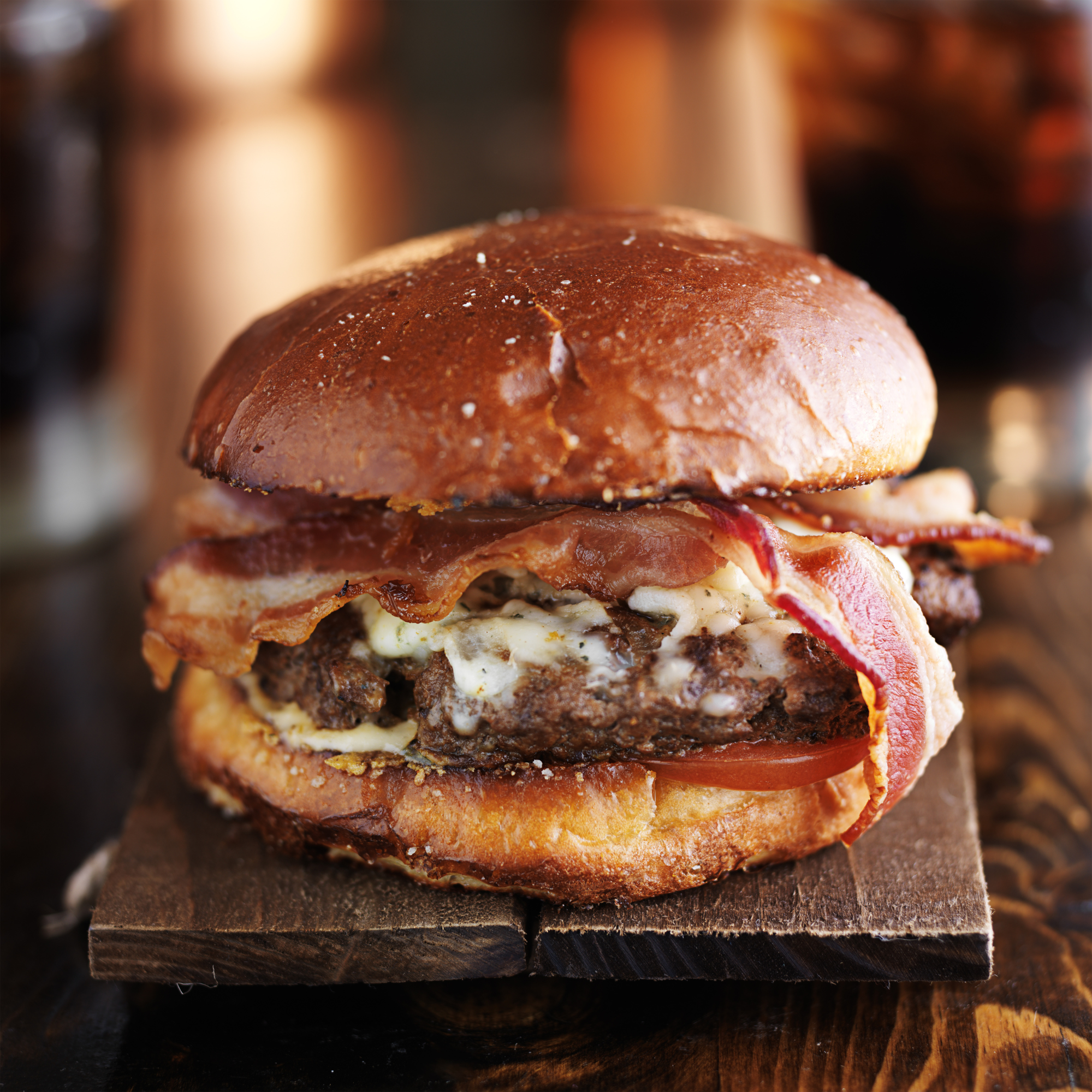 A close-up of a gourmet double cheeseburger on a dark wooden board, featuring a glossy toasted bun, melted cheese, bacon, and a beef patty on a dark wooden table.