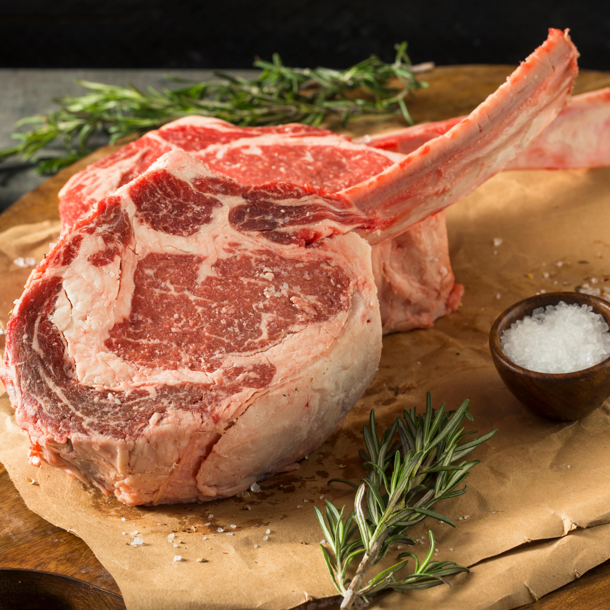 Two raw, thick-cut USDA Prime Black Angus bone-in ribeye steaks seasoned with sea salt and fresh rosemary on butcher paper, processed direct by Winston Cattle Co.